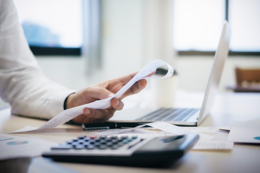 Open laptop and a calculator on a desk with a man's arm holding a piece of paper right in front of the laptop