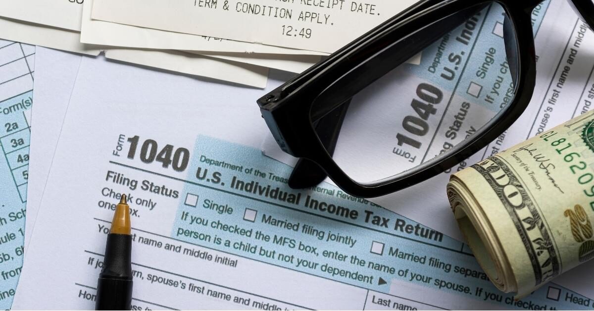 Scattered tax papers with glasses, pen, and rolled bills on top of a 1040 form