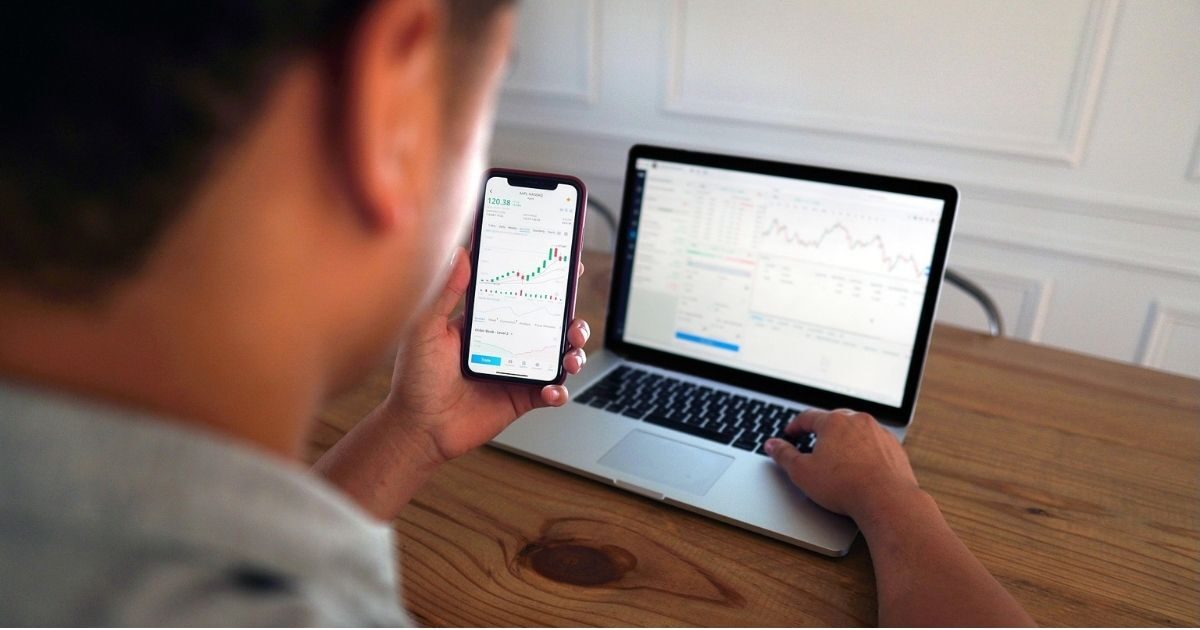 Man sitting at a desk holding a phone with the screen on stocks. A laptop is open on the desk with the man's hand on it with the screen showing stocks